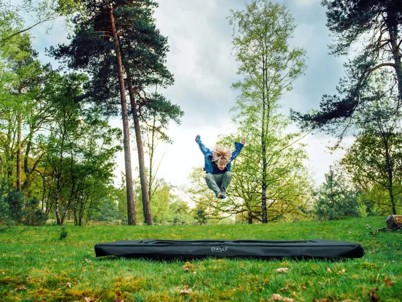 Kind springt hoog boven een rechthoekige Etan-trampoline in een bosrijke omgeving